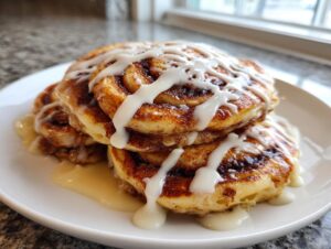 A stack of three fluffy cinnamon roll pancakes drizzled generously with white cream cheese icing.