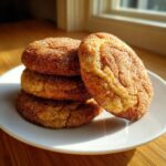 A stack of four soft, thick churro cookies coated heavily in cinnamon sugar, resting on a white plate.