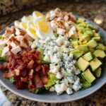 A beautifully arranged classic cobb salad featuring rows of grilled chicken, hard-boiled eggs, bacon, blue cheese, and avocado over lettuce.