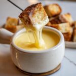 A piece of toasted bread is dipped into a bowl of creamy, melted cheese fondue recipe, showing long cheese pulls.