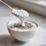 A spoonful of creamy chia seed pudding is dripping back into a white bowl filled with the mixture.