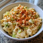 Close-up of creamy deviled egg pasta salad with celery, topped with paprika and herbs in a white bowl.