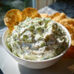 A bowl of creamy dill pickle dip topped with fresh dill and served with potato chips.