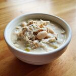A close-up of a white bowl filled with creamy white chicken chili, featuring shredded chicken and white beans.