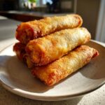 A stack of four golden brown, crispy fried egg roll appetizers resting on a light ceramic plate.