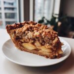 A close-up of a thick slice of dutch apple pie recipe served on a white plate, showing spiced apple filling and a crumb topping.