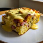 A square slice of eggs benedict casserole topped generously with creamy hollandaise sauce and parsley.