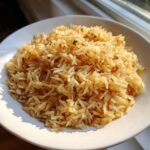 A close-up of fluffy, golden brown rice pilaf seasoned with herbs served in a white bowl.