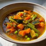 Close-up of a rich, savory vegetable soup featuring chunks of carrots, zucchini, celery, and bright green peas.