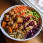 Close-up of a colorful buddha bowl featuring quinoa, roasted sweet potatoes, shredded carrots, edamame, and a rich peanut dressing.
