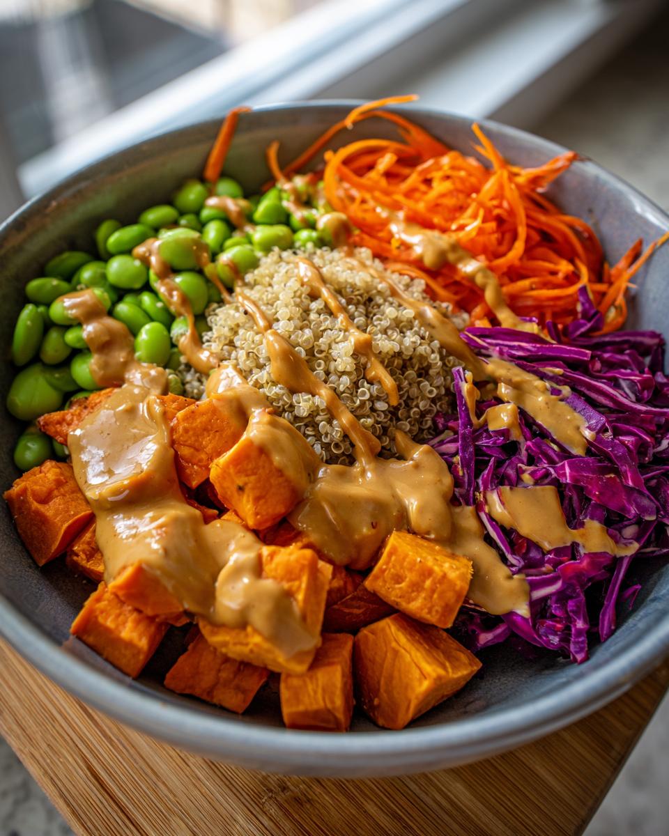 Close-up of a colorful buddha bowl featuring quinoa, roasted sweet potatoes, edamame, shredded carrots, and purple cabbage with peanut sauce.