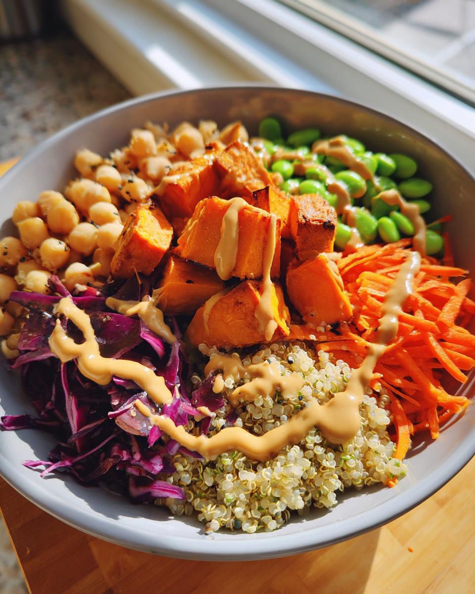 Close-up of a vibrant buddha bowl featuring quinoa, roasted sweet potato, chickpeas, edamame, shredded carrots, and red cabbage with a peanut dressing.