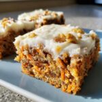 A close-up view of a thick slice of carrot cake bars topped with cream cheese frosting and walnuts.