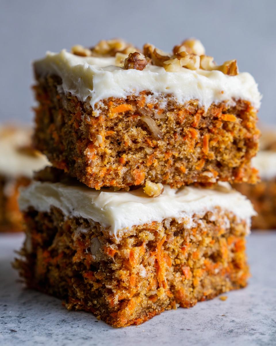 Two stacked squares of incredible carrot cake bars topped with thick cream cheese frosting and chopped walnuts.
