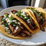 Close-up of three delicious steak tacos filled with sliced marinated meat, topped with fresh diced white onion and cilantro.