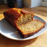 A golden-brown loaf of moist banana bread recipe, with one thick slice cut and resting beside it on a plate.