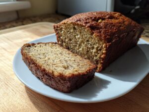 A freshly baked loaf of banana bread recipe, with one thick slice cut and resting beside it on a white plate.