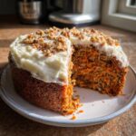 A slice removed from a homemade carrot cake topped with thick cream cheese frosting and chopped walnuts.