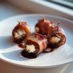 Four bacon wrapped stuffed dates filled with white cheese, drizzled with glaze on a white plate.