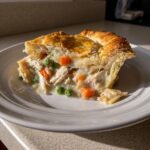 A close-up of a flaky slice of chicken pot pie showing shredded chicken, carrots, and peas in a creamy sauce.