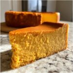 Close-up of a thick slice of rich, orange pumpkin cheesecake with the rest of the pumpkin cheesecake blurred in the background.