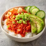 A close-up of a spicy crab sushi bowl featuring rice topped with bright red spicy crab mix, avocado, kimchi, and scallions.