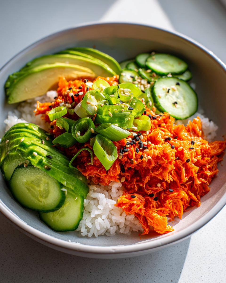 A close-up of a spicy crab sushi bowl featuring white rice topped with bright orange spicy crab mix, sliced avocado, cucumbers, and green onions.