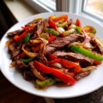 A close-up of perfectly cooked steak fajitas featuring sliced steak, red and green bell peppers, and onions on a white plate.