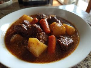 Close-up of a steaming white bowl filled with rich, dark brown beef stew, featuring chunks of tender beef, potatoes, and carrots.