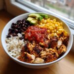 A white bowl filled with a colorful chicken burrito bowl featuring seasoned chicken, rice, black beans, corn, salsa, and avocado slices.