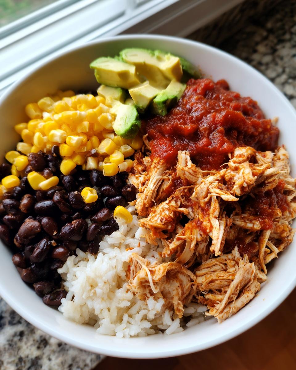 A white bowl filled with a delicious chicken burrito bowl featuring shredded chicken, rice, black beans, corn, salsa, and avocado slices.