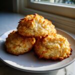 A stack of four golden brown, crispy-edged coconut cookies resting on a light gray plate near a window.