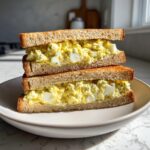 Two halves of a thick egg salad sandwich stacked, showing creamy filling with visible egg chunks on toasted bread.