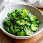 A close-up of bright green, thinly sliced Asian cucumber salad tossed in a savory dressing and topped with toasted sesame seeds.