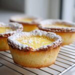 A close-up of one of the freshly baked baby lemon impossible pies dusted with powdered sugar, cooling on a wire rack.