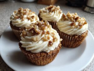Four perfectly frosted banana walnut cupcakes topped with a generous sprinkle of chopped walnuts, resting on a white plate.