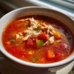Close-up of a bowl of bright red maryland crab soup topped with shredded white meat and diced vegetables.