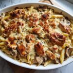 Close-up of a baked chicken marsala casserole featuring penne pasta, mushrooms, and browned chicken pieces topped with melted cheese.