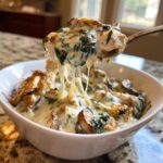 A spoonful of cheesy chicken spinach mushroom low carb oven dish being lifted from a white bowl, showing melted cheese pull.