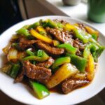 A close-up, appetizing photo of Chinese pepper steak featuring tender beef strips mixed with bright green and yellow bell peppers in a savory brown sauce.