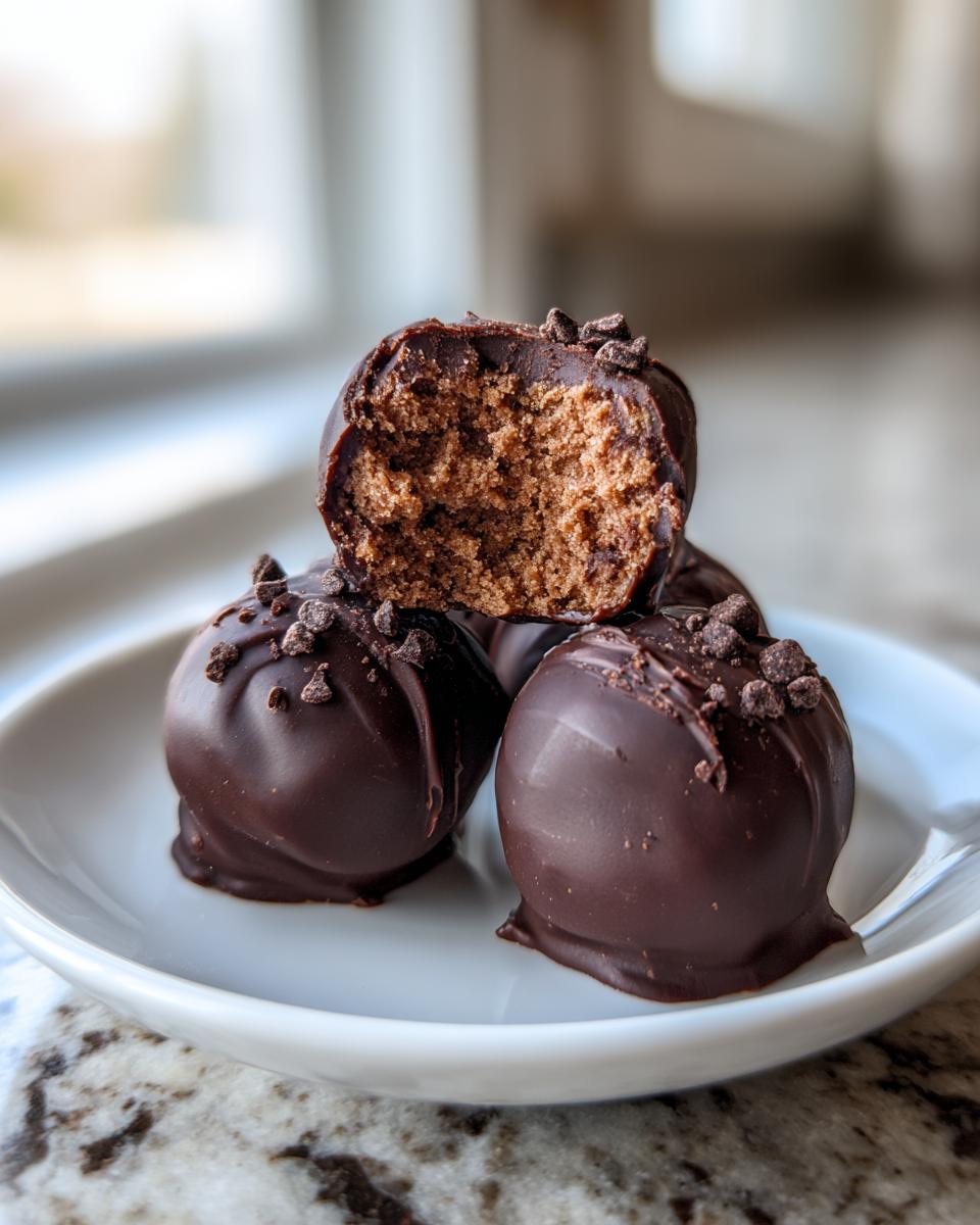A stack of rich chocolate chip cookie dough truffles, one bitten to show the soft interior, topped with mini chocolate chips.
