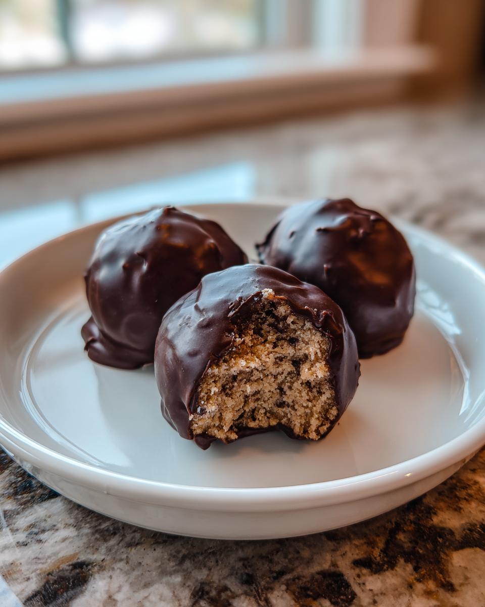 Three chocolate chip cookie dough truffles on a white plate, one is bitten showing the dough interior.