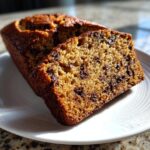 A close-up of sliced chocolate chip vegan banana bread resting on a white plate, showing a moist, golden crumb.