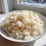 A white bowl filled with fluffy coconut ginger garlic rice, illuminated by bright sunlight near a window.