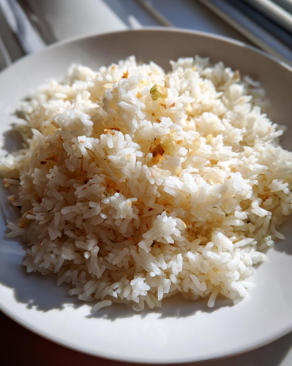 A close-up of fluffy white rice, prepared as a coconut ginger garlic rice recipe, served on a white plate under bright sunlight.