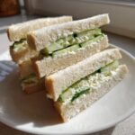 Four triangles of classic cucumber sandwiches stacked on a white plate, showing creamy filling and fresh cucumber slices.
