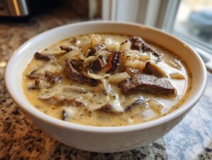 A close-up of a hearty bowl of philly cheese steak soup, featuring beef strips, mushrooms, and caramelized onions in a creamy broth.