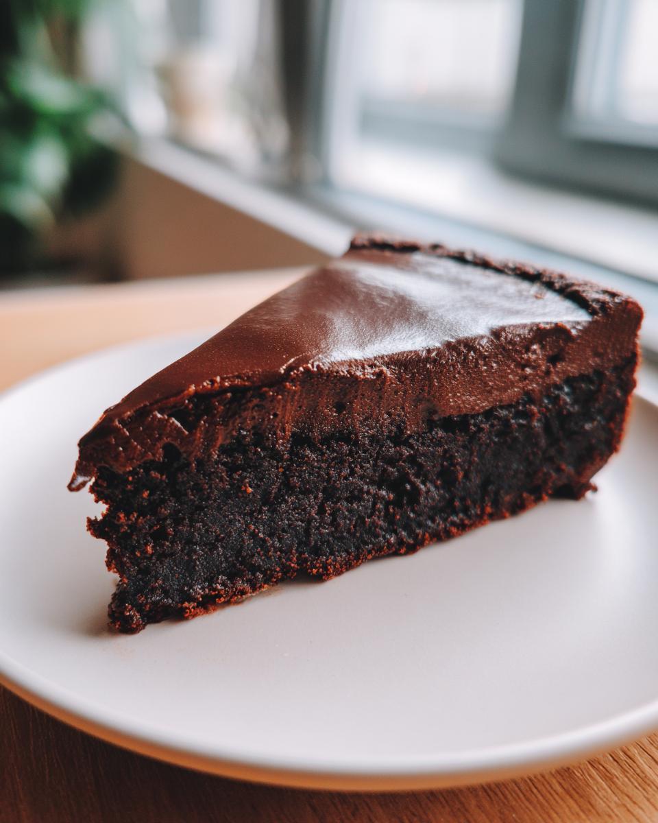 A decadent slice of flourless chocolate cake with a thick, glossy chocolate topping served on a white plate.