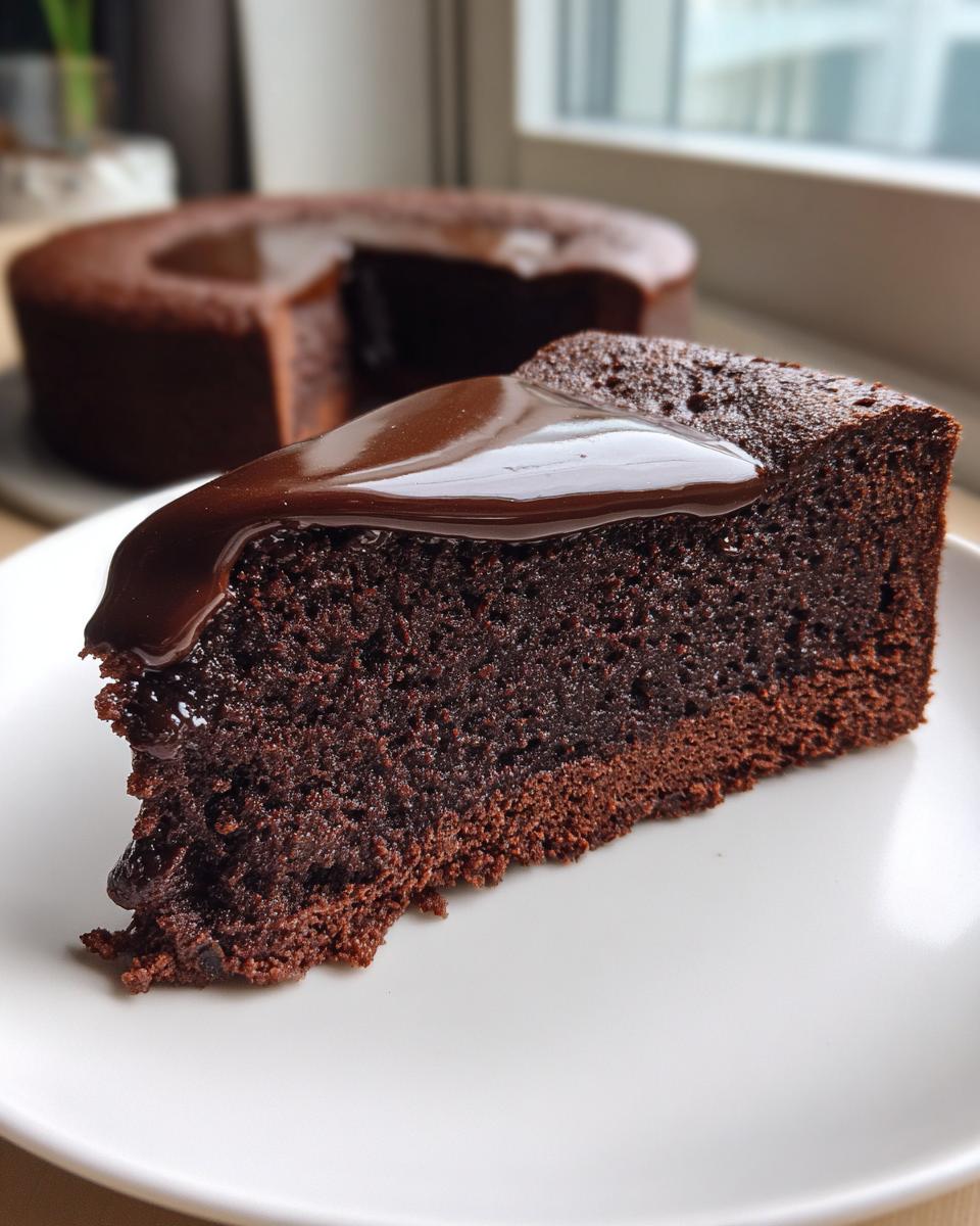 A close-up slice of moist flourless chocolate cake topped with glossy chocolate ganache, with the rest of the cake blurred in the background.