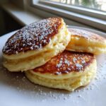 Close-up of three thick, fluffy buttermilk pancakes stacked on a white plate, topped with powdered sugar.